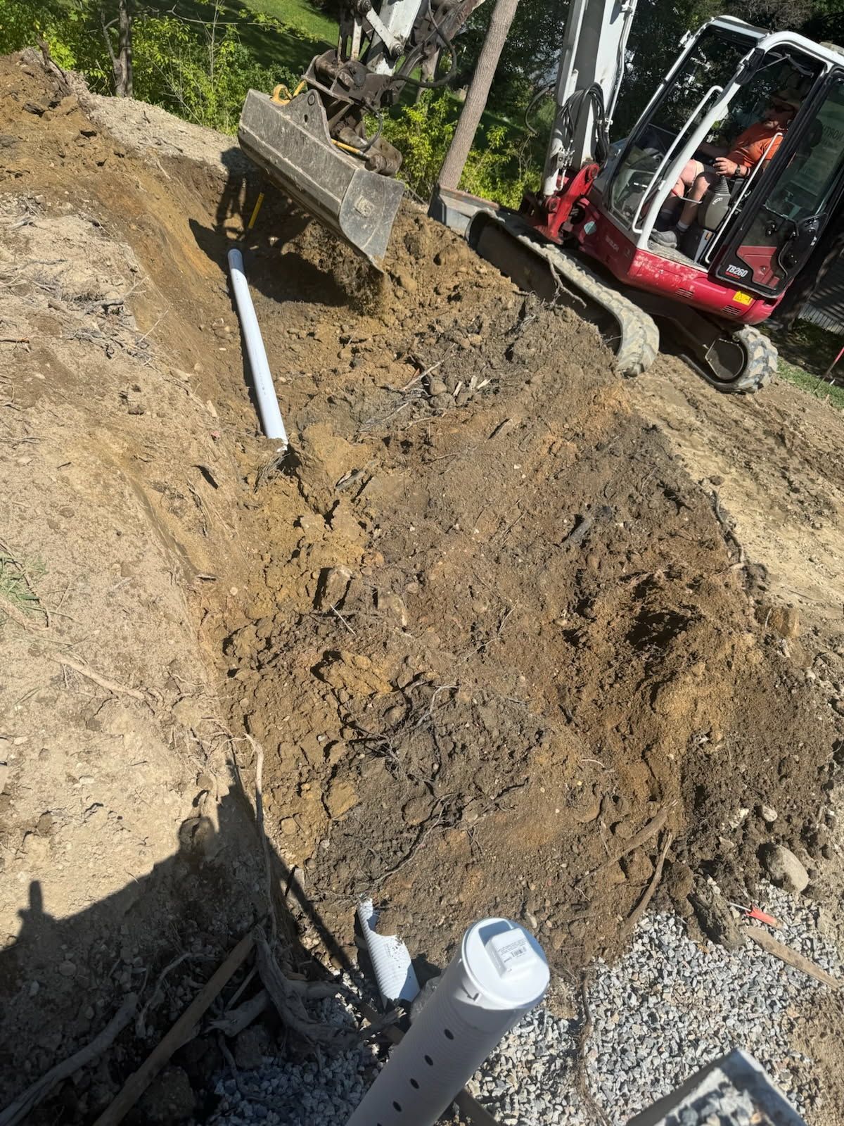 Excavator digging a trench beside a gravel path, with soil piled around the hole.