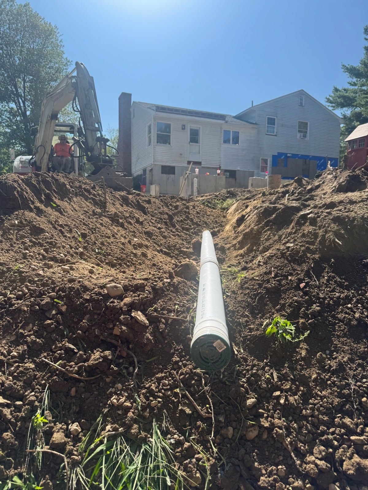 Large white pipe in a dirt trench below a house, with an excavator nearby under a clear blue sky