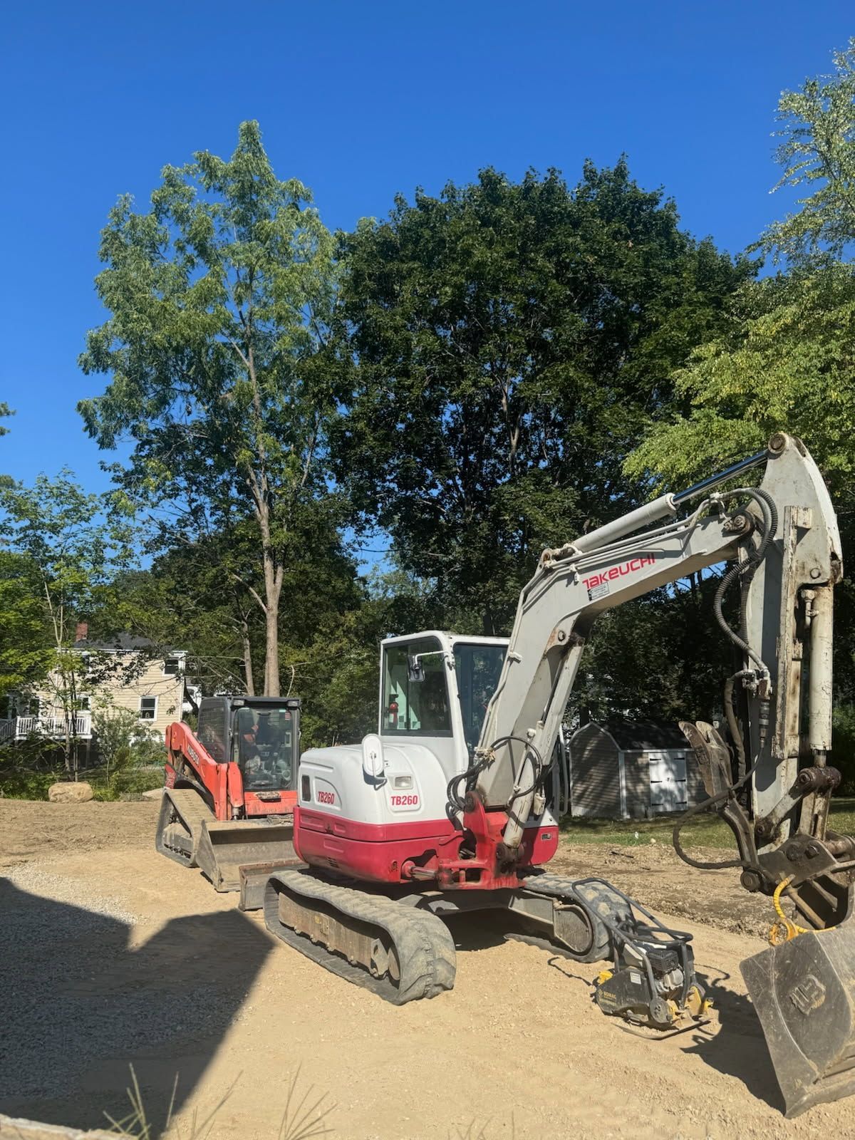 Red and white excavator parked on a dirt path under trees on a sunny day