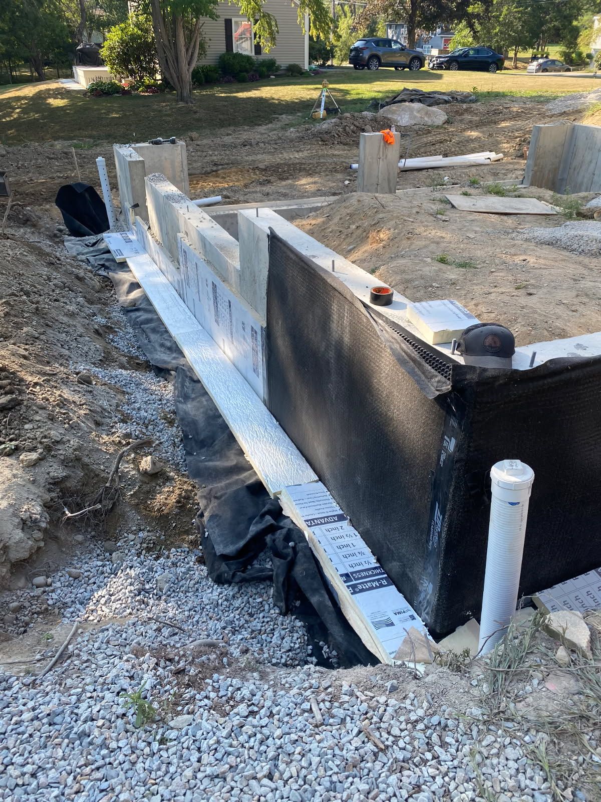 Concrete foundation wall under construction with black waterproofing, gravel, and white PVC pipes.