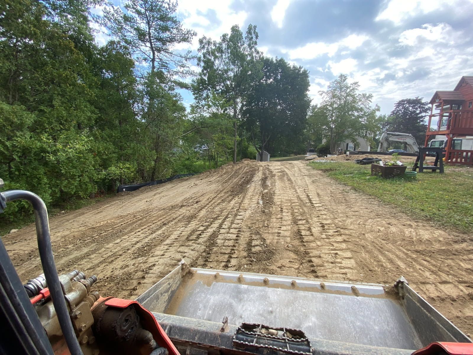 Freshly graded dirt lot beside trees and a red building, viewed from a construction vehicle