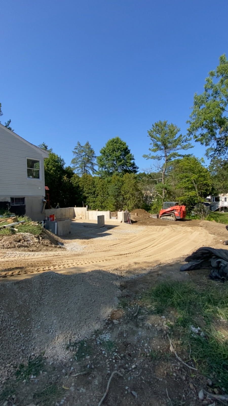 New house construction site with dirt driveway, trees, and a small orange excavator under a clear blue sky