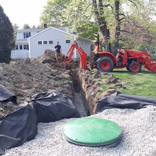 Backhoe trenching beside a house with black tarp and gravel-covered septic tank lid in the foreground