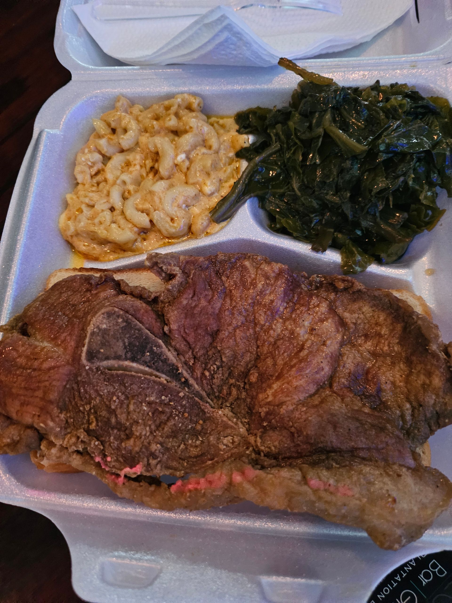A styrofoam container filled with meat , macaroni and cheese , and greens.