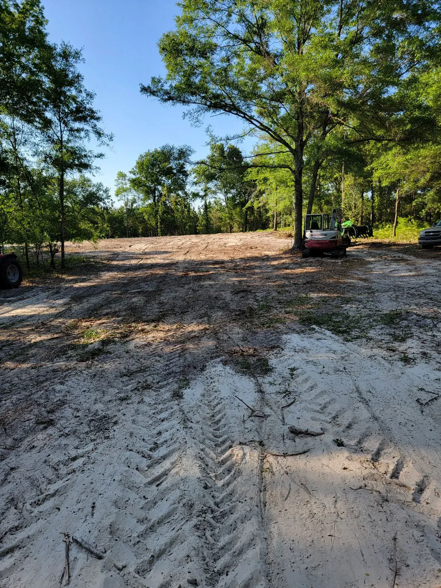 Sandy clearing with tire tracks, surrounded by trees. Cars parked on the right.