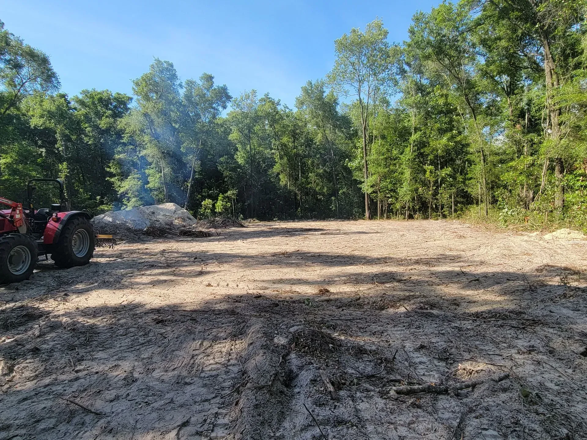 Tractor clearing land with debris pile burning in the background, surrounded by trees.