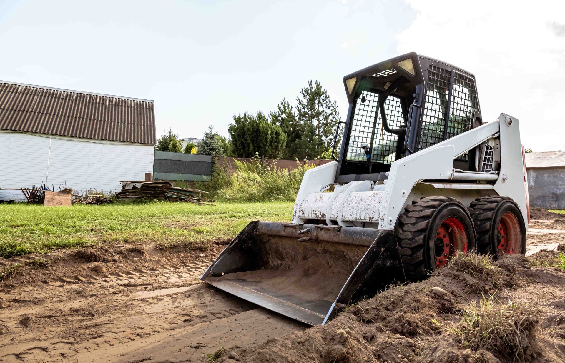 White skid-steer loader with front bucket working in a dirt field near a barn.