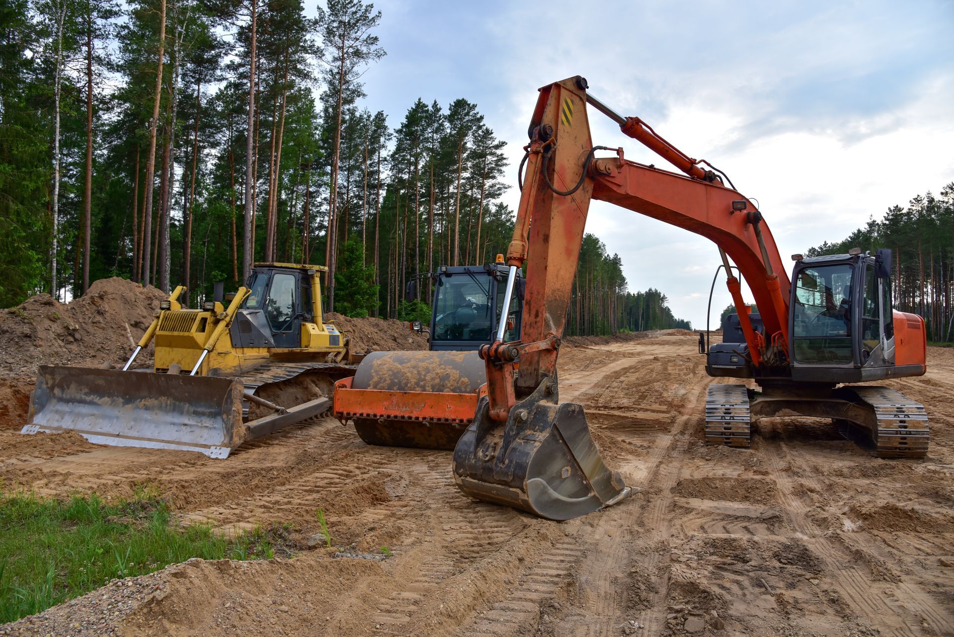 Three construction machines on a dirt road in a forest clearing, including a yellow bulldozer and orange excavator.