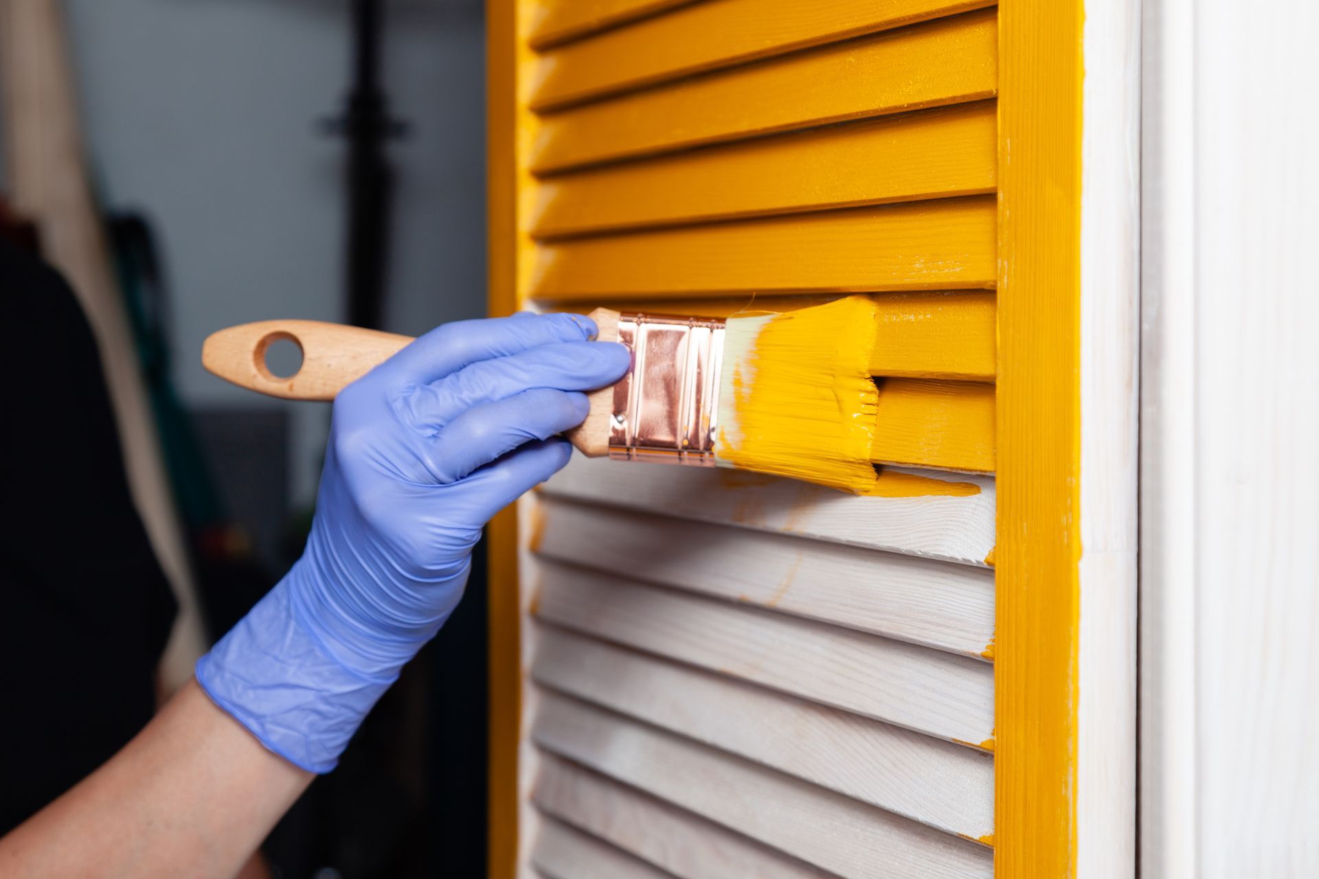 Person wearing blue gloves painting yellow shutters with a paintbrush.