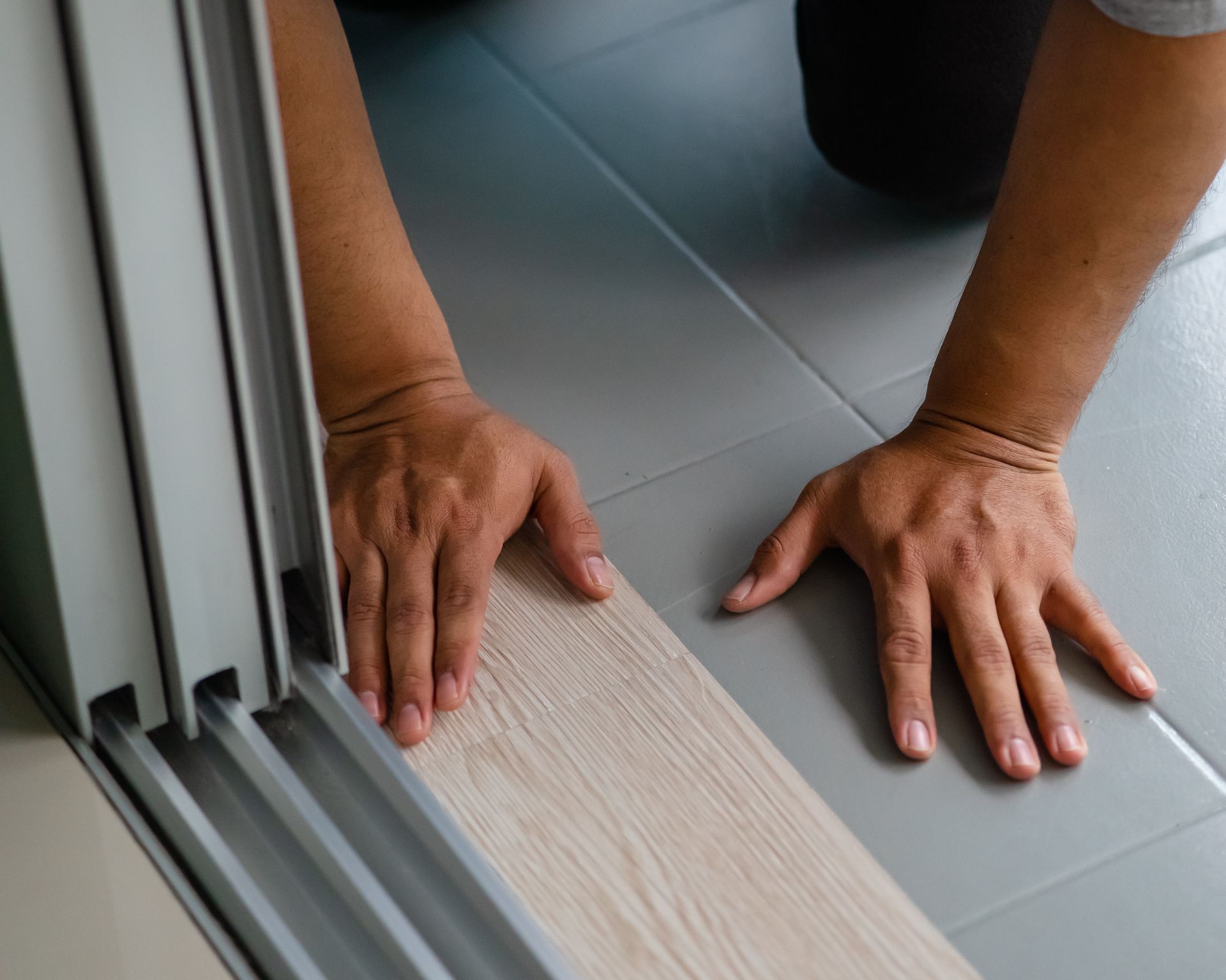 Person installing wood-look flooring next to a sliding door, hands close to the floor.