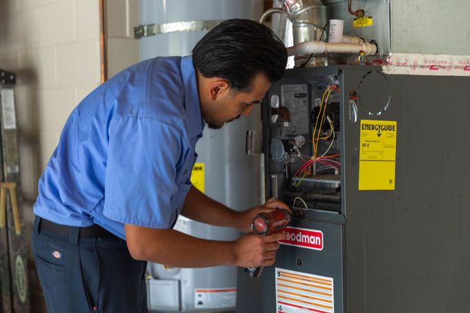 Technician wearing a blue uniform uses a drill to repair a furnace unit in a utility room.