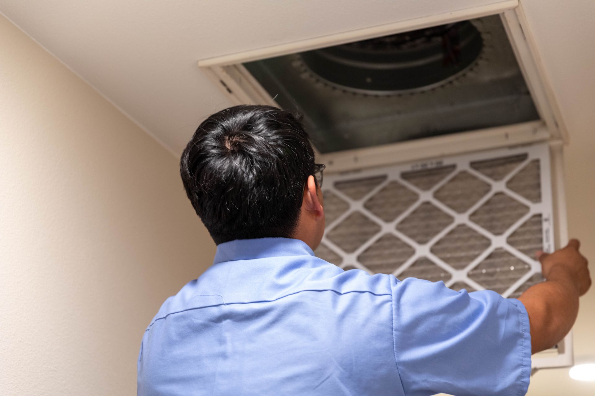A person in a blue shirt stands on a ladder, using a yellow cloth to clean a square vent on a white ceiling.