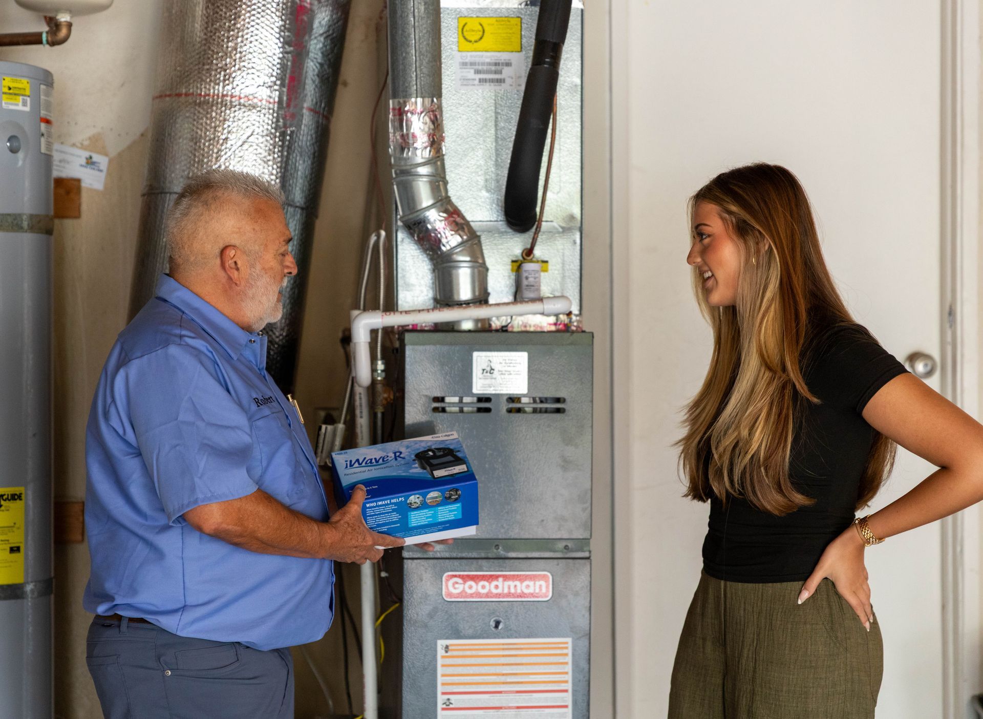 Two professionals in blue work shirts inspect a furnace and water heater setup in a utility room.