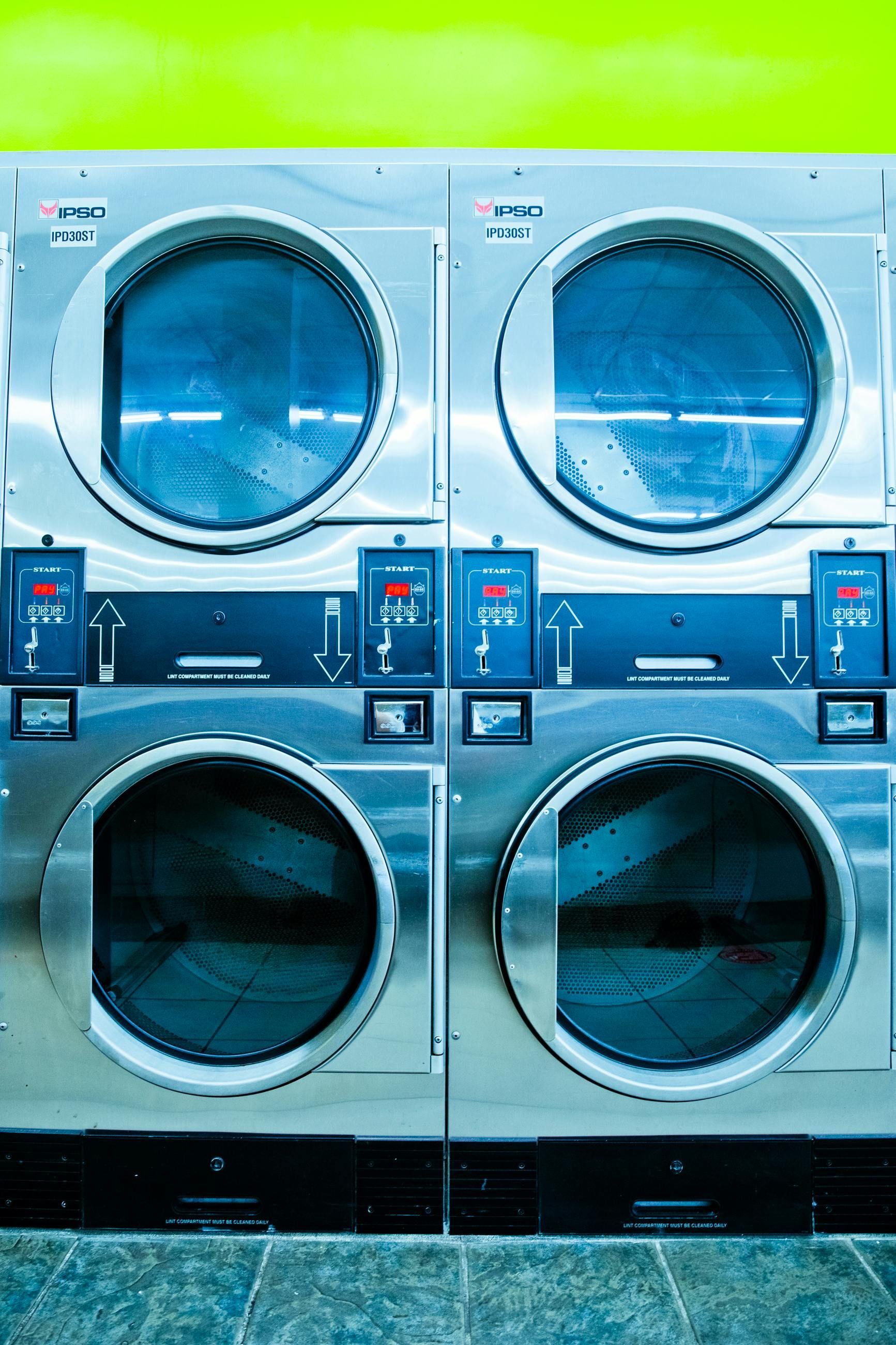 A person in a blue button-down shirt cleaning a dryer vent hose with a vacuum attachment in a laundry room.