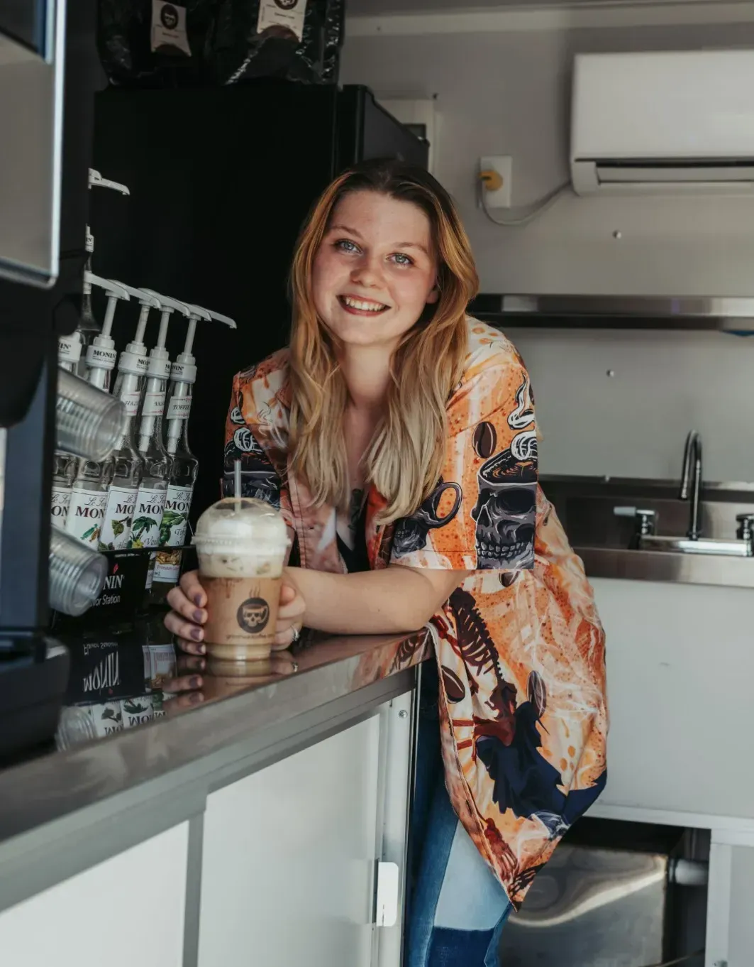 Woman smiling behind a counter holding a iced drink in a small kitchen area
