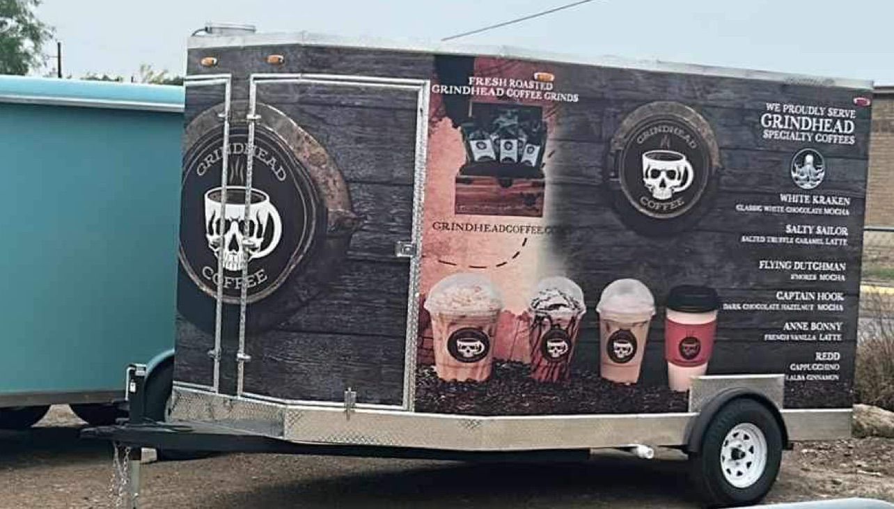 Black food trailer with coffee cup graphics and menu text parked beside a blue tanker truck