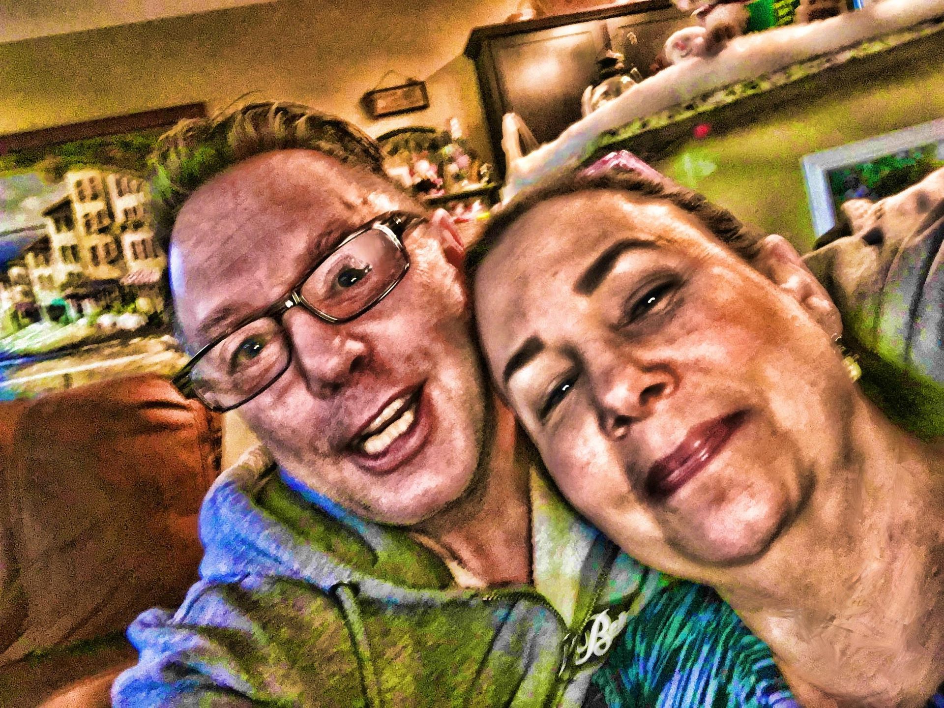 Two smiling people taking a selfie indoors, one wearing glasses and a blue jacket
