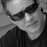 Close-up black-and-white portrait of a person wearing sunglasses and a dark shirt, looking slightly downward.