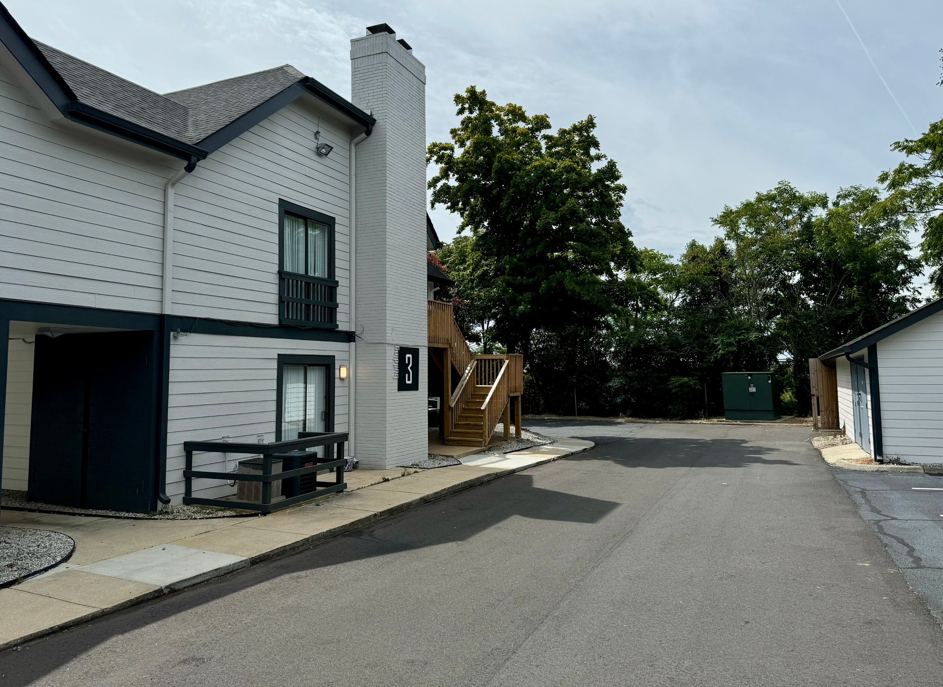 White brick building beside a paved driveway with a stairway and trees in the background