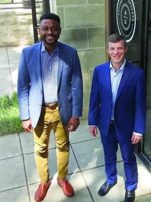Two men in suits stand outside a building. One wears a blue blazer and yellow pants, the other a blue suit.
