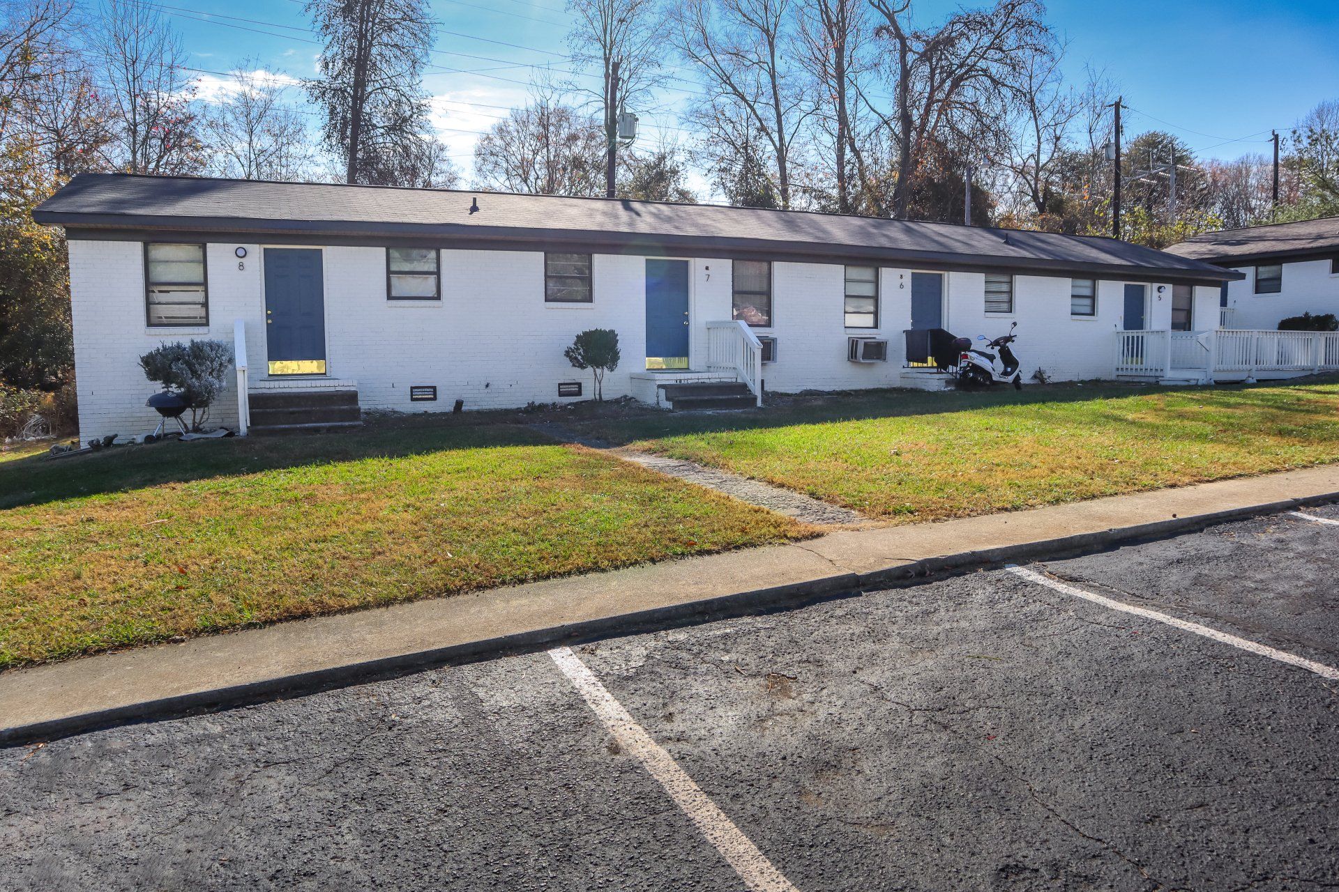 Row of white apartments with blue doors and black roof; parking lot in front, grass in between.