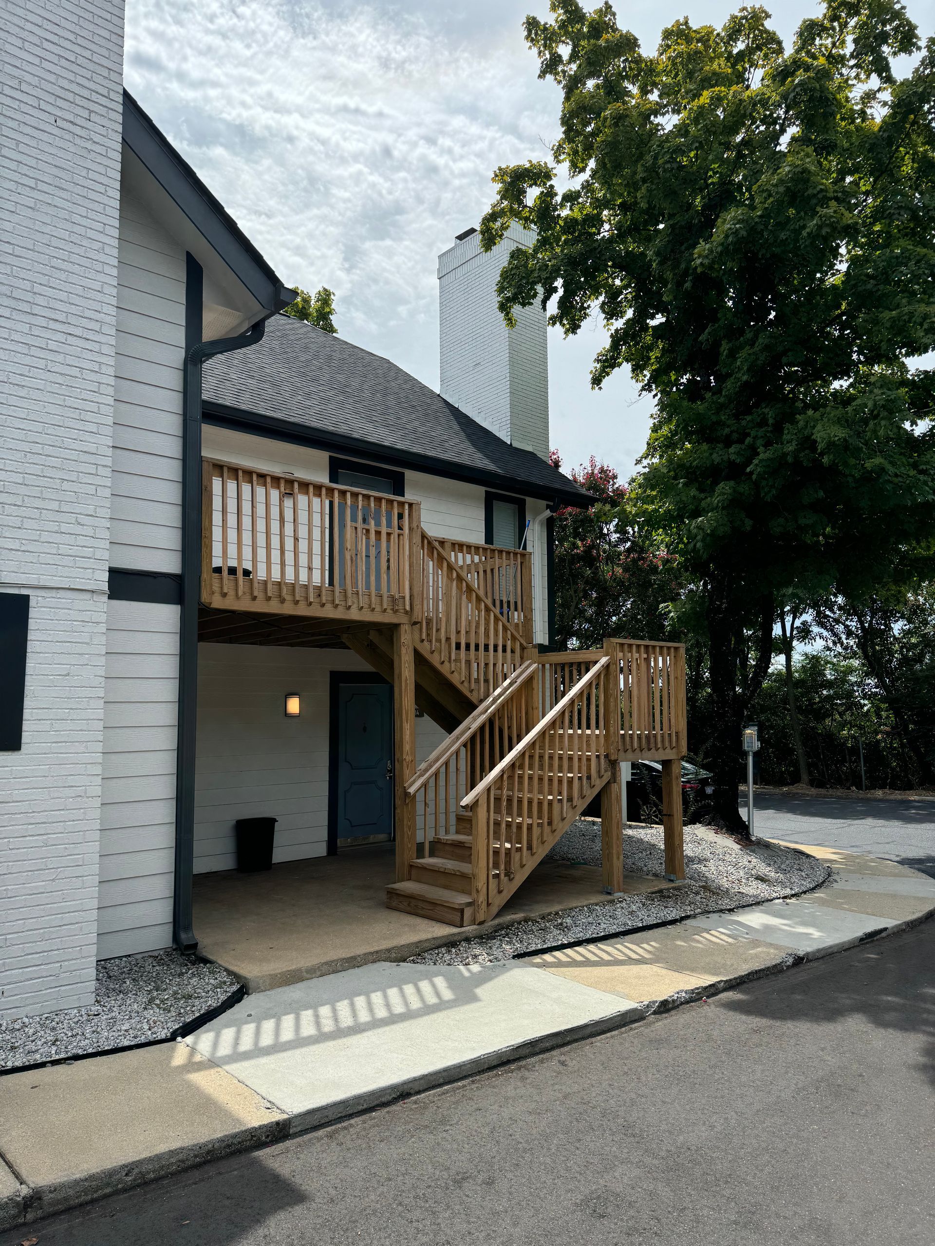 Exterior of a white brick house with a wooden staircase and balcony under a large tree