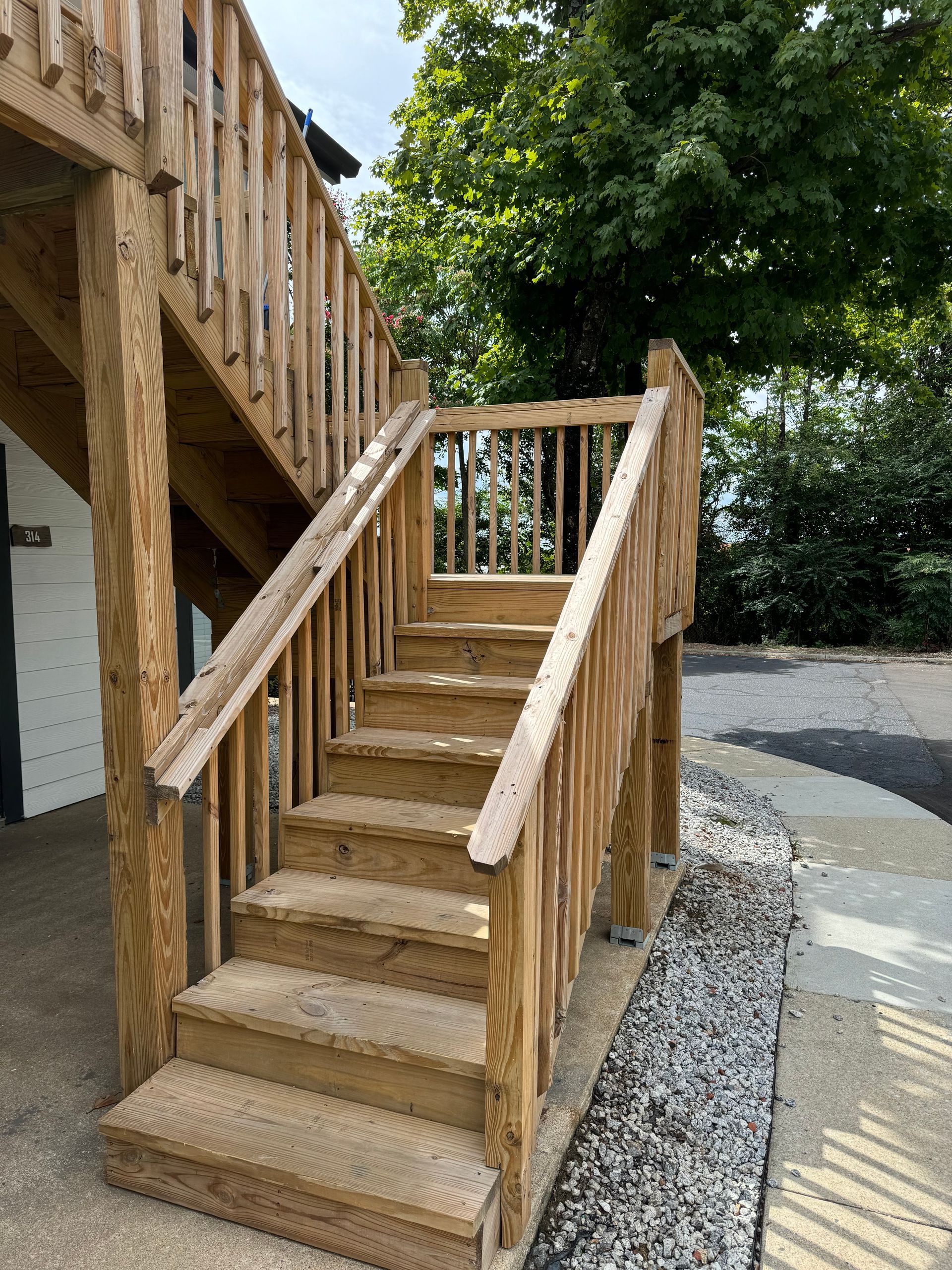 Wooden outdoor staircase with railings beside a building, bordered by gravel and a large tree.
