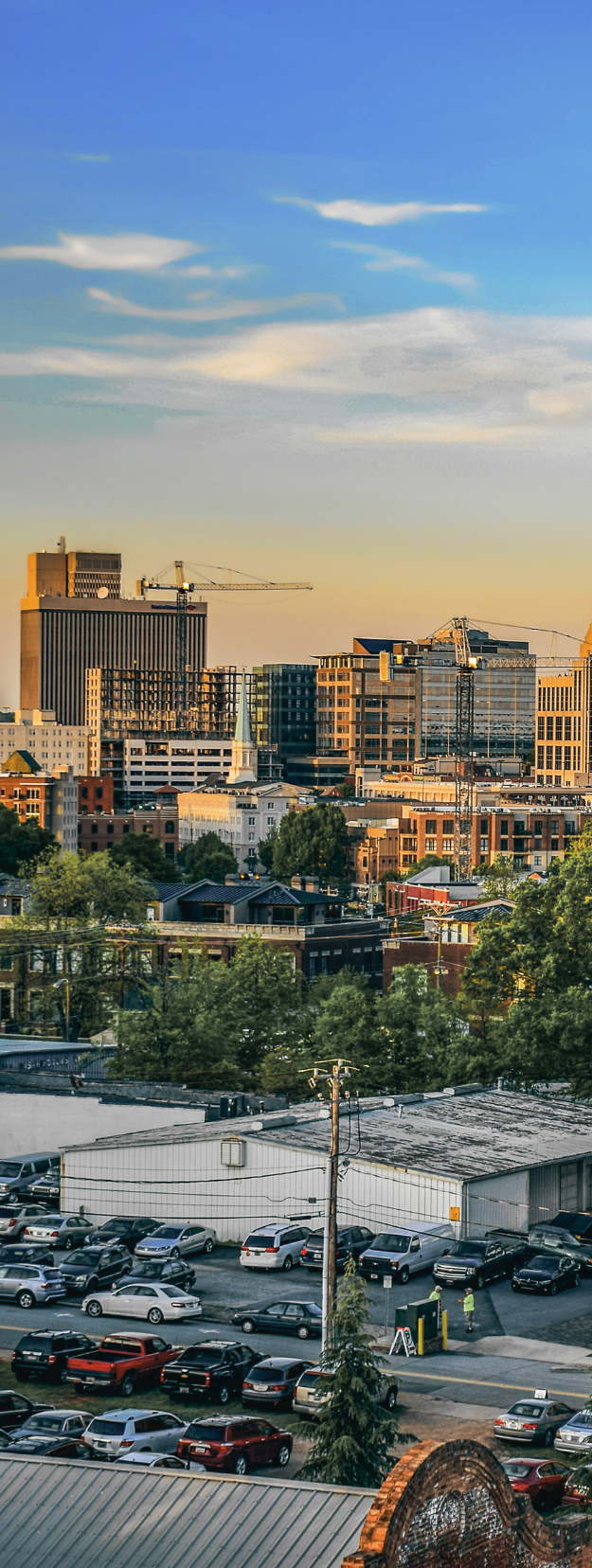 City skyline at dusk with a mix of buildings, trees, and parked cars. Blue and yellow sky.