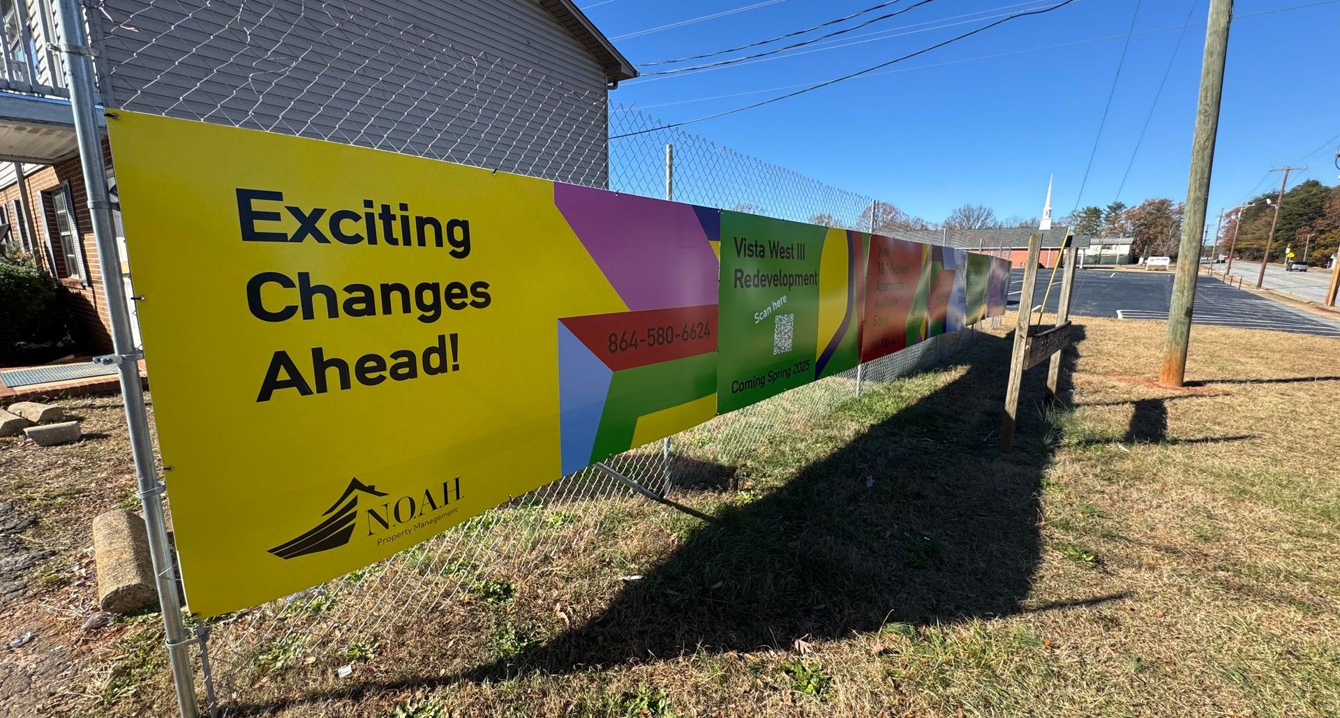 Yard sign reading “Exciting Changes Ahead!” on a fence beside a sidewalk and street.