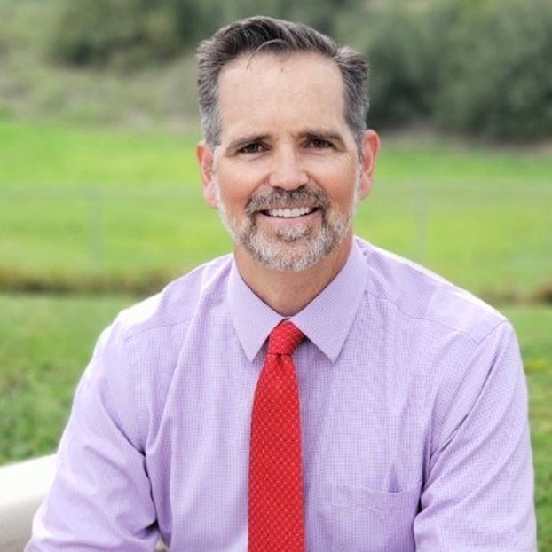 Smiling man in a lavender shirt and red tie seated outdoors with a blurred green background