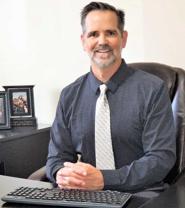 Man sitting at a desk with a keyboard, wearing a dark shirt and tie, smiling in an office.