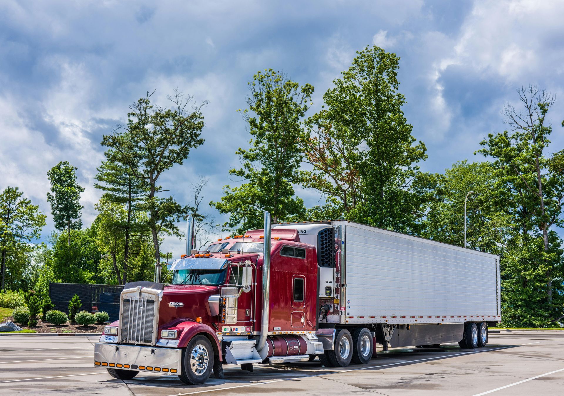 Red semi-truck with silver trailer parked in a lot under a blue sky and green trees.