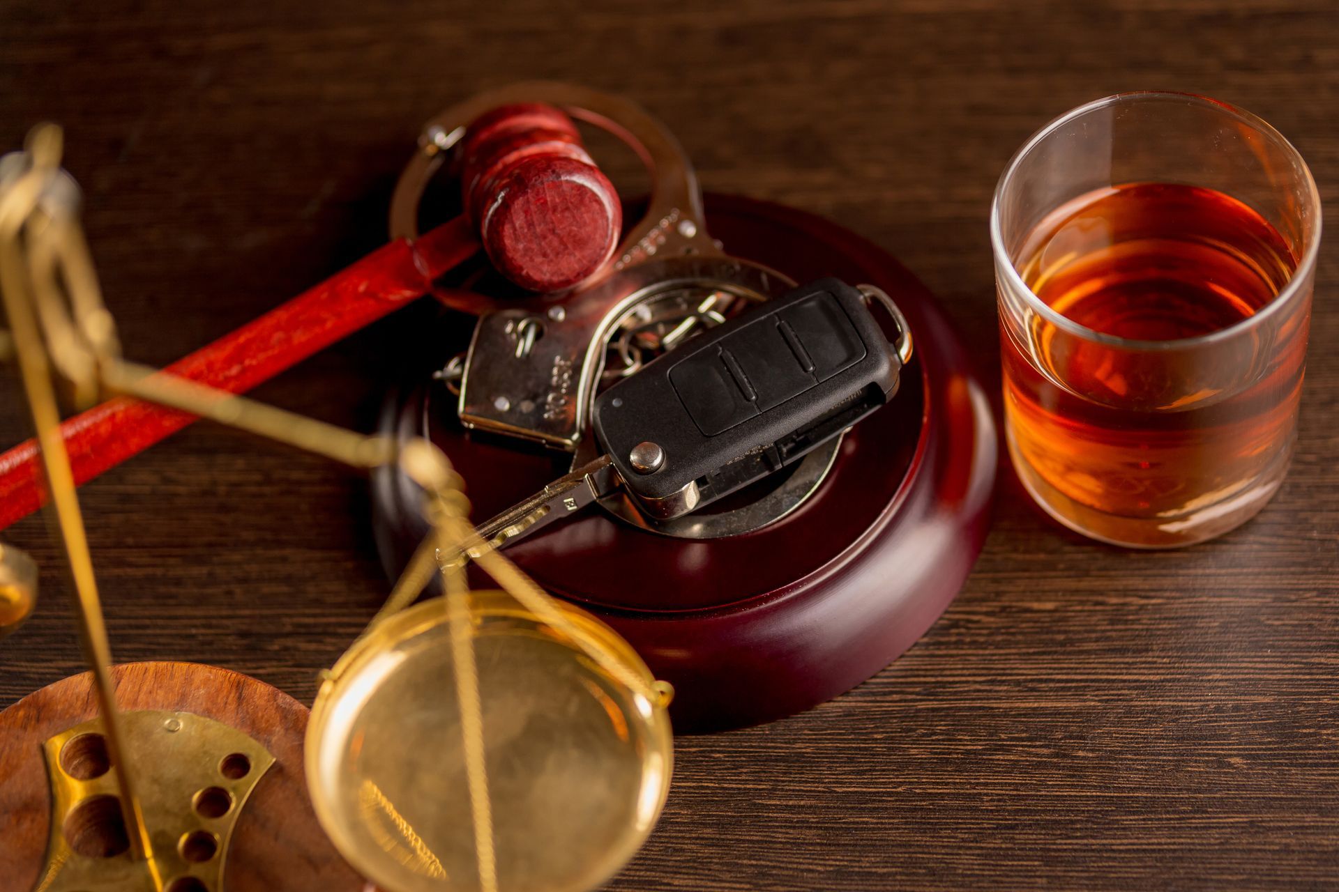 Scales of justice, gavel, handcuffs, car keys, and a drink on a wooden surface. Scales of justice, gavel, handcuffs, car keys, and a drink on a wooden surface.
