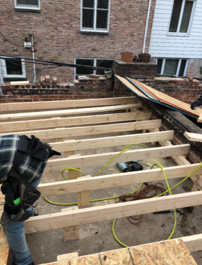 A man is working on a wooden deck in front of a brick building.