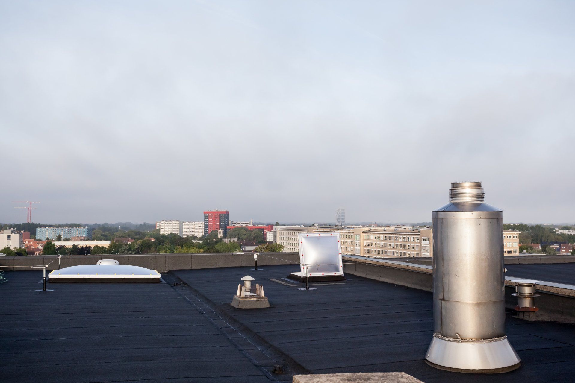 A flat black roof with ventilation pipes and skylights overlooking a city landscape under a cloudy sky.