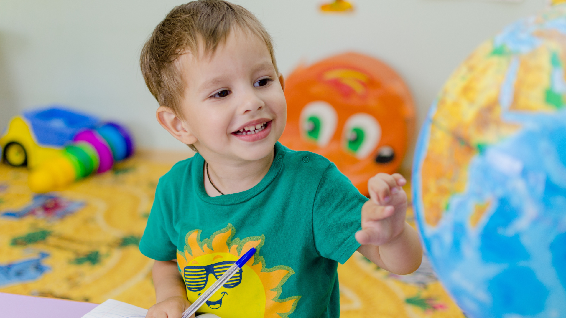 Smiling child in a green shirt sitting on a bed with colorful toys and large painted balls behind them
