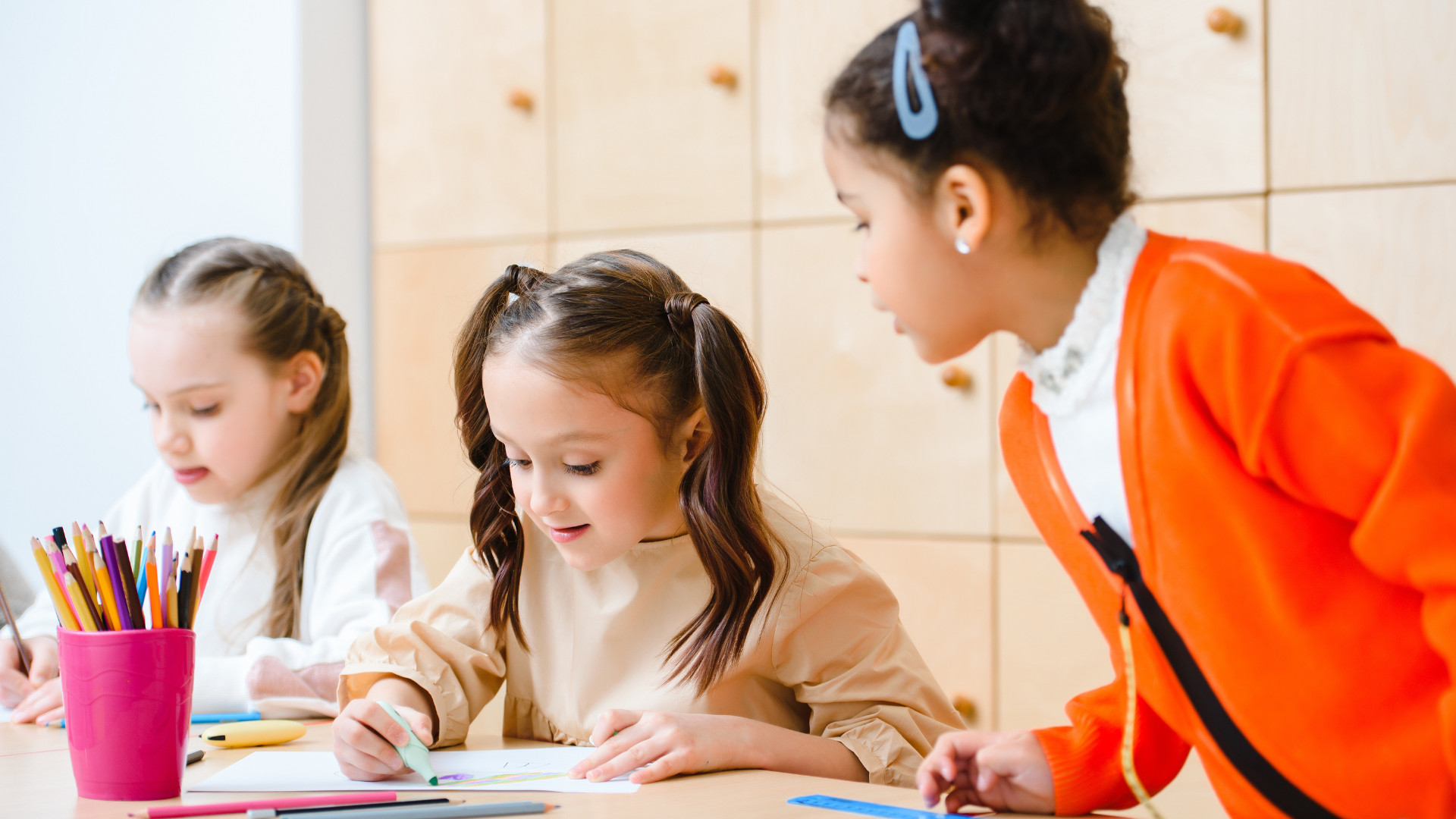 Children coloring at a table while a teacher leans over to help in a classroom