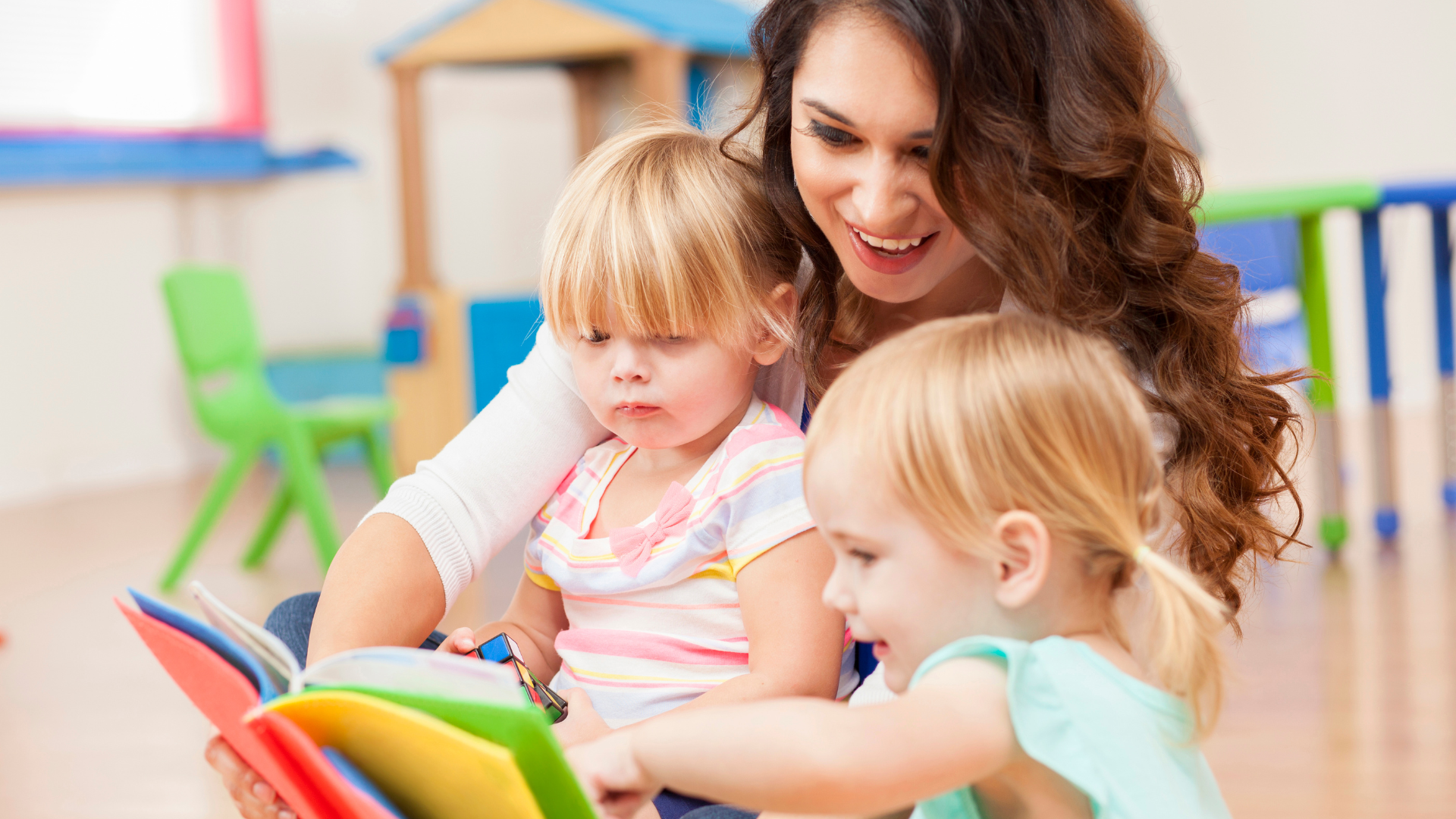 Caregiver reading a colorful book with two small children in a bright classroom