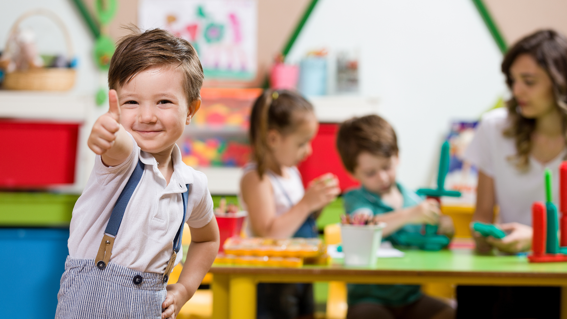 Child smiling and giving a thumbs-up in a colorful classroom, with other children playing at tables behind him.