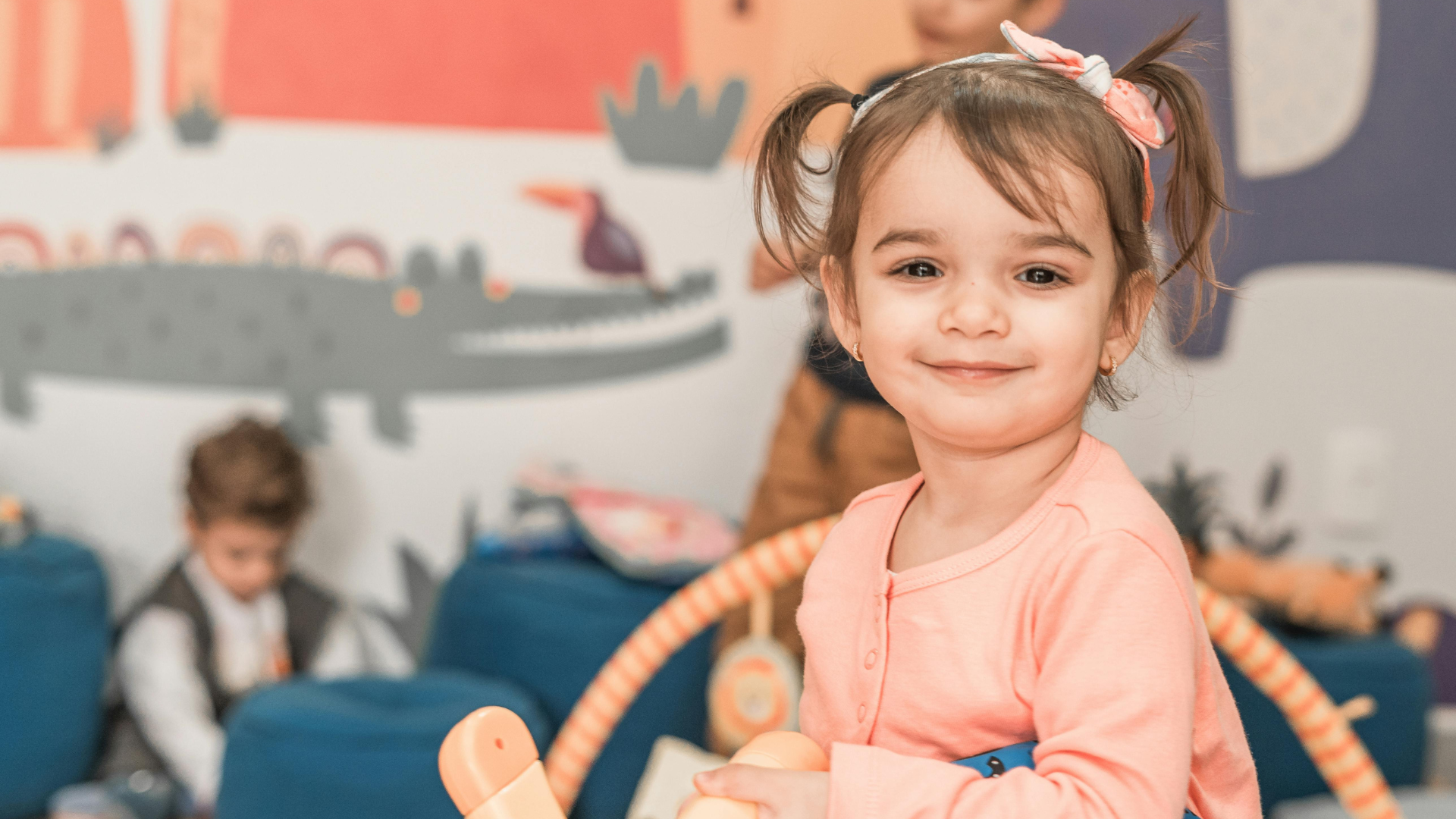 Smiling child in a pink shirt sitting in a playroom with toys and other children in the background