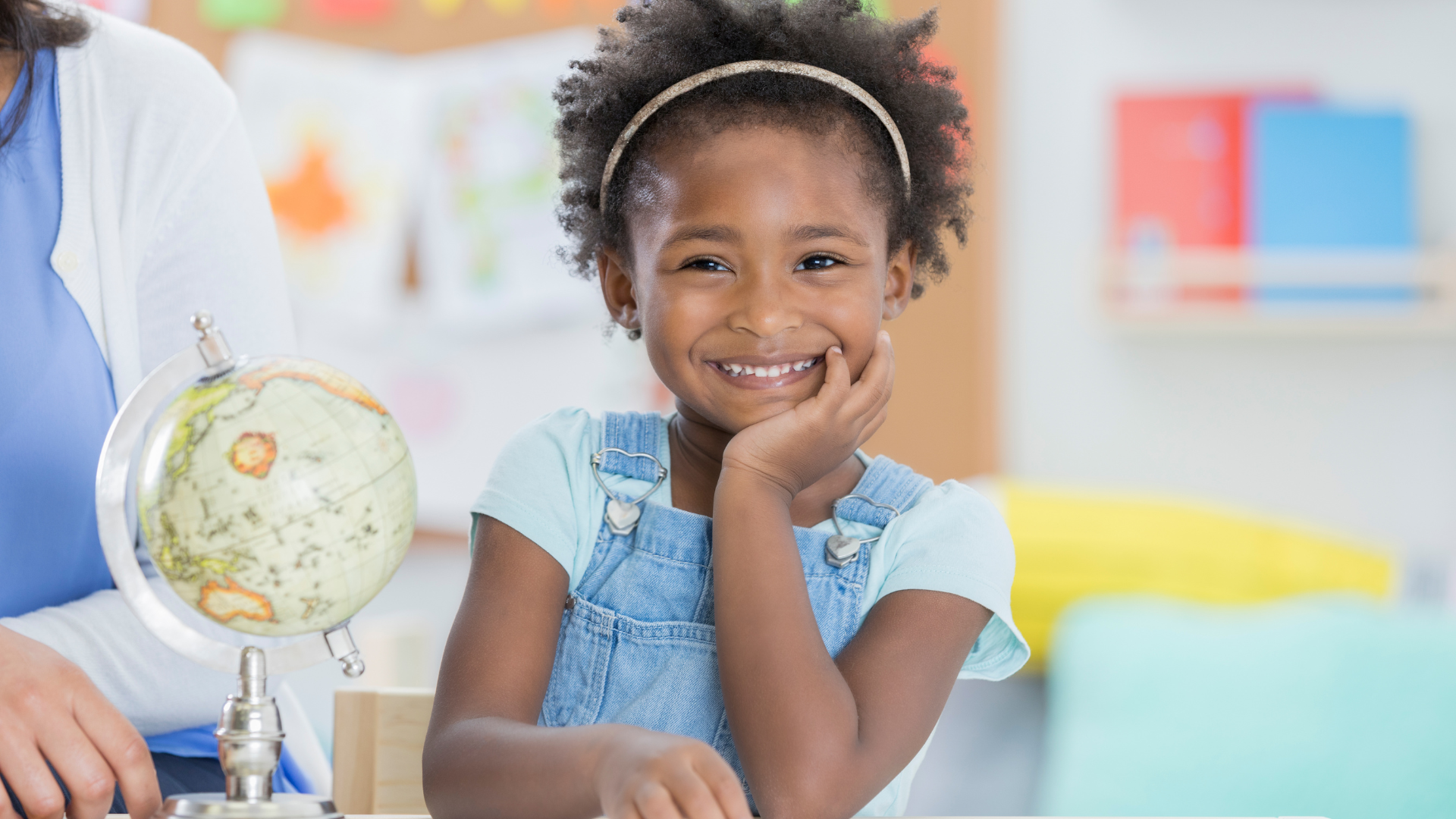 Smiling child in a light blue shirt beside a globe in a bright classroom
