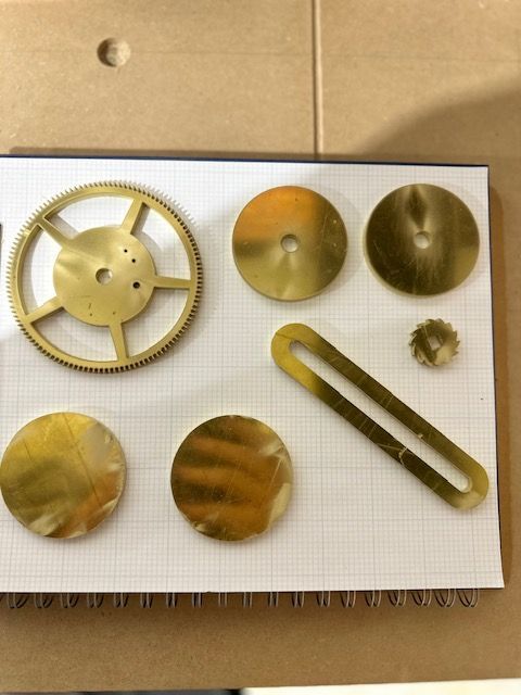 Assorted brass-colored metal parts and discs laid out on a white foam board on a cardboard surface.