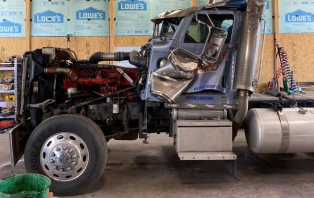 Damaged semi-truck cab in a workshop, showing significant body damage and exposed engine components.