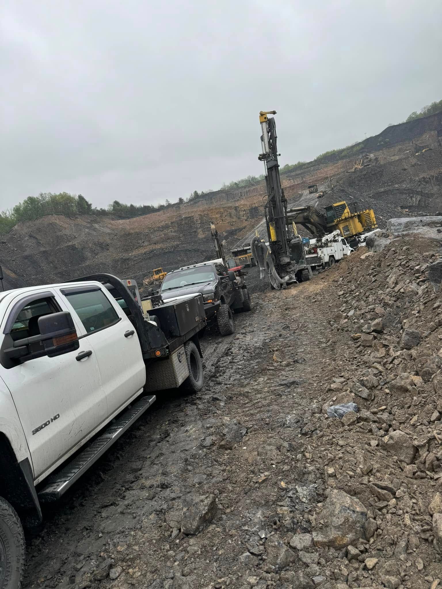 White truck towing a drill rig on a muddy, rocky road near a hillside. Cloudy day.