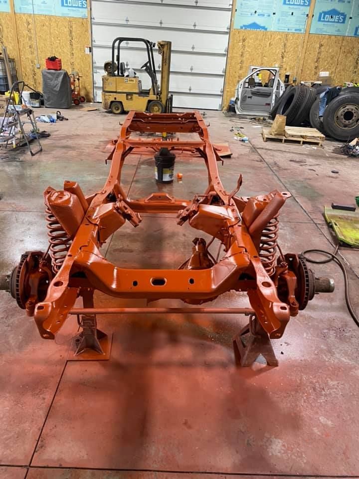 Orange-painted car frame on jack stands in a garage. A forklift and other parts are visible in the background.