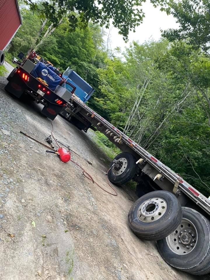 Flatbed semi-truck angled on a sloped gravel driveway. Welding equipment and trees are visible.