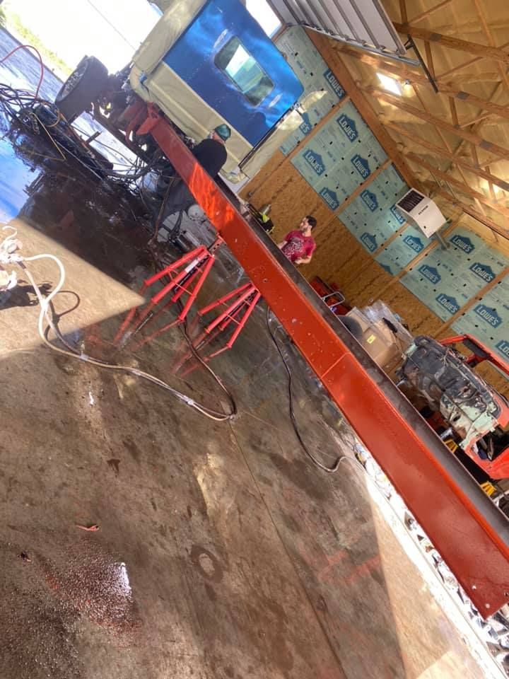 A red truck bed elevated in a workshop. A person stands by a beam. Concrete floor.