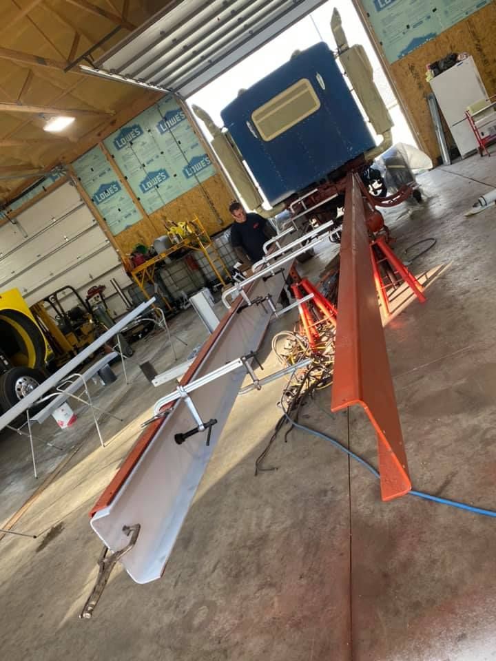 Man working on orange and white vehicle component in a garage, truck in the background.