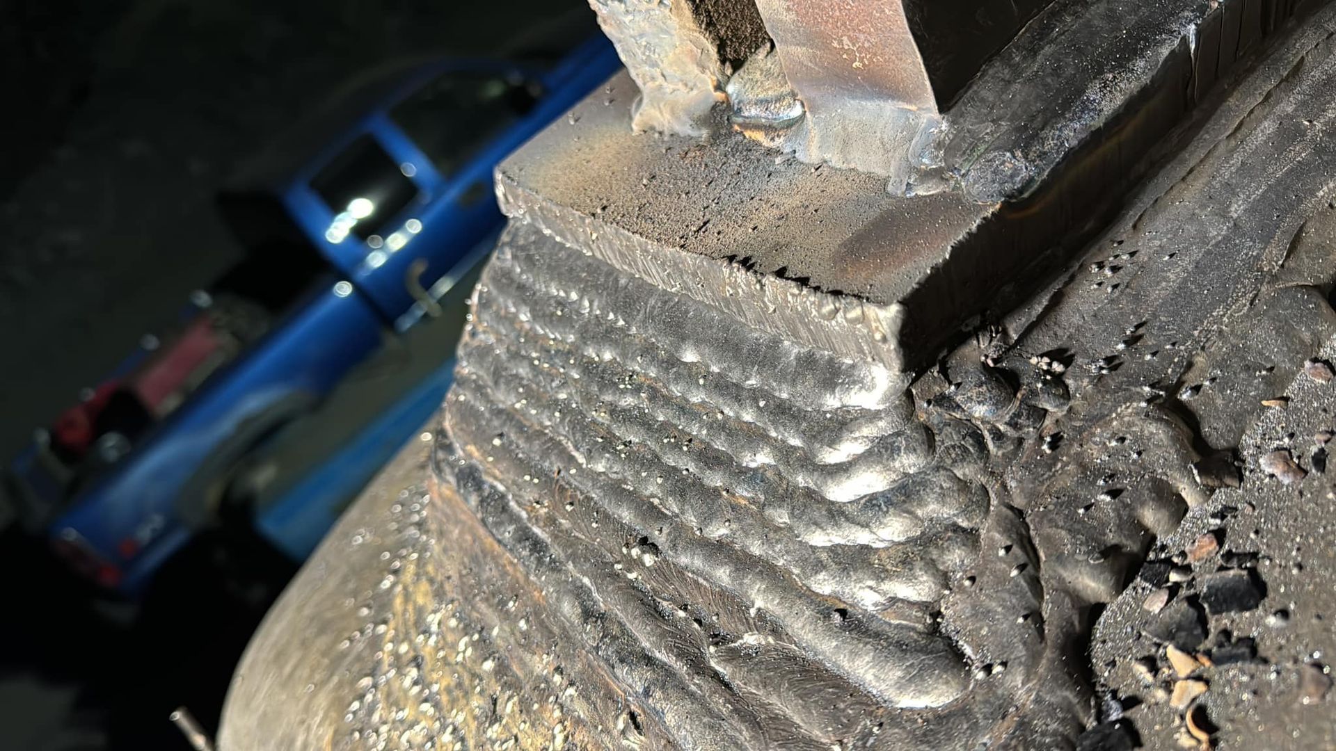 Welded metal structure: Layered weld beads connecting a block to a curved surface, set against a blurred backdrop of a blue pickup truck.