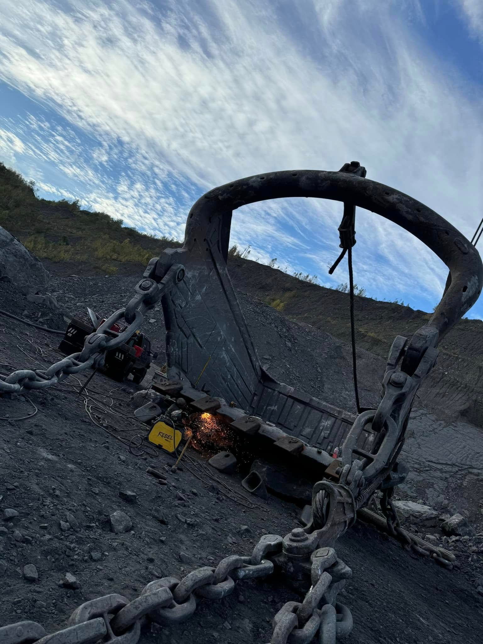 Welder repairs a giant dragline bucket in a coal mine. Blue sky with clouds overhead.